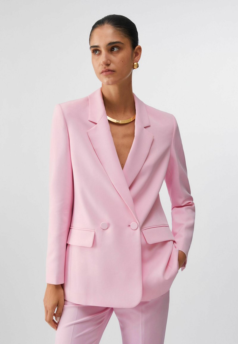 Femme aux cheveux foncés portant un blazer rose à double boutonnage et un pantalon assorti, accessoirisée avec des boucles d'oreilles et un collier en or, sur un fond blanc.