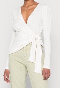 Top bianco testurizzato con maniche lunghe e scollatura a V profonda, con dettaglio annodato in vita. Abbinato a pantaloni verde chiaro.