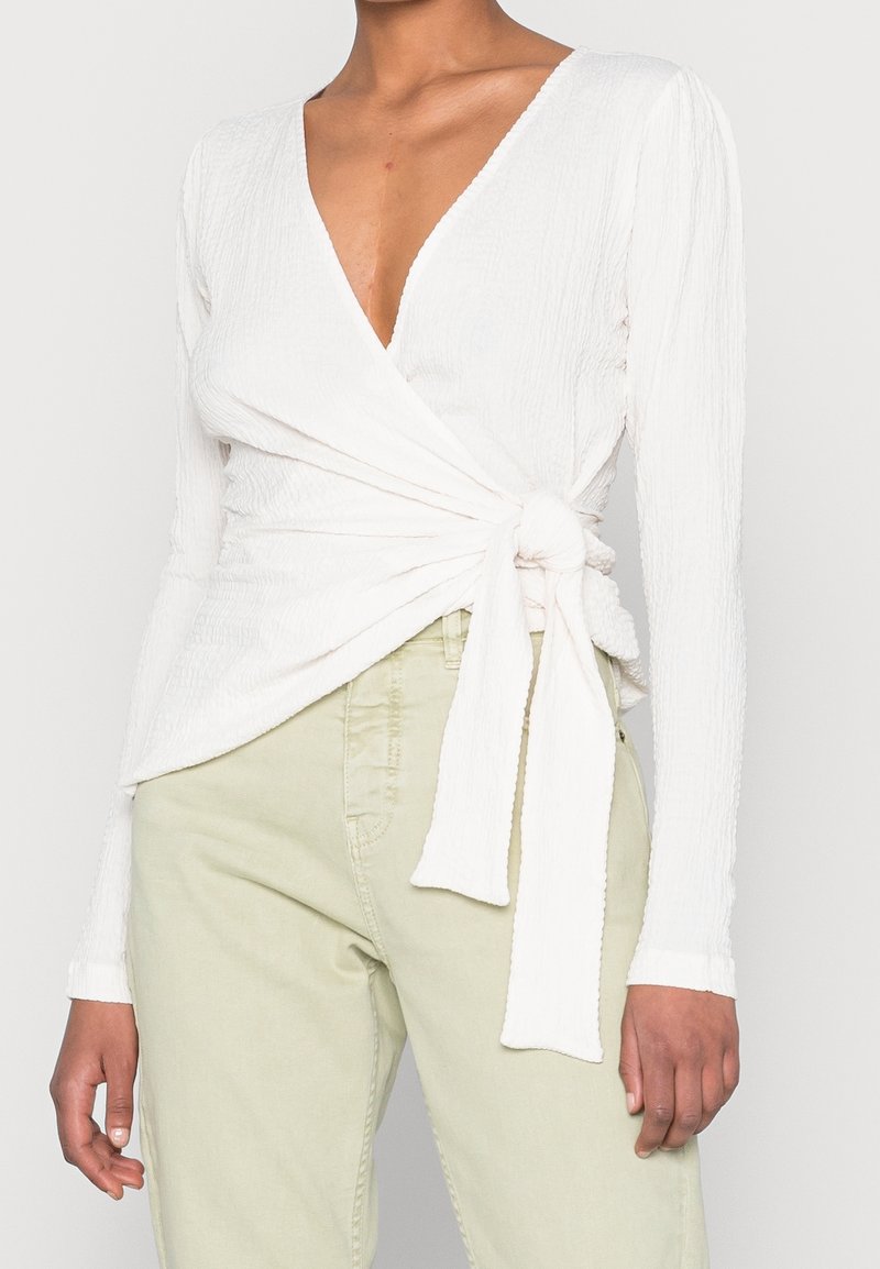 Top bianco testurizzato con maniche lunghe e scollatura a V profonda, con dettaglio annodato in vita. Abbinato a pantaloni verde chiaro.