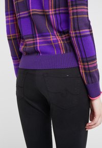 Suéter a cuadros púrpura y negro con puños y bajo acanalados, combinado con jeans negros que presentan costuras decorativas en los bolsillos traseros.