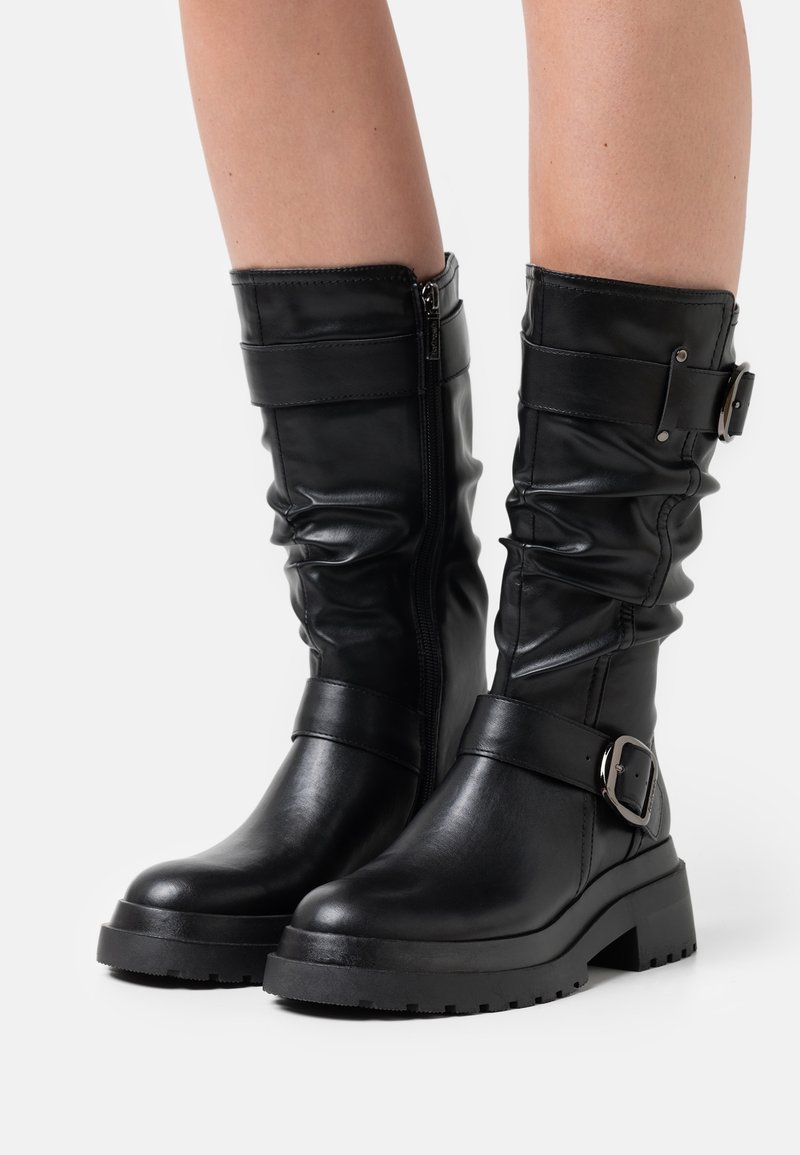 Czarne skórzane botki do kostki z grubymi podeszwami, bocznymi zamkami błyskawicznymi i dekoracyjnymi paskami z metalowymi klamrami, noszone na gołych nogach.