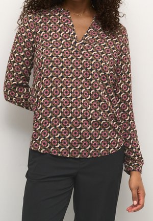 Vrouw draagt een lange mouwen blouse met een circulair geometrisch patroon in roze, beige en zwart, gecombineerd met effen zwarte broek tegen een witte achtergrond.