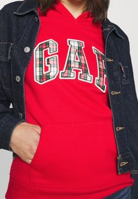 Červená mikina s kapucí s károvaným nápisem „GAP“, vrstvená pod tmavě modrou džínovou bundou s knoflíky a vyšíváním. Viditelná je kapsa mikiny.
