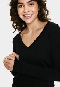 Fekete, hosszú ujjú felső V-nyakkal és hullámos szegéllyel. Puha kötött anyag és testre szabott dizájn, amelyen összekulcsolt kezek láthatók.