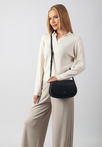 Black leather shoulder bag with a curved shape and flap closure, featuring a gold accent on the strap. Model wears a cream sweater and beige trousers.