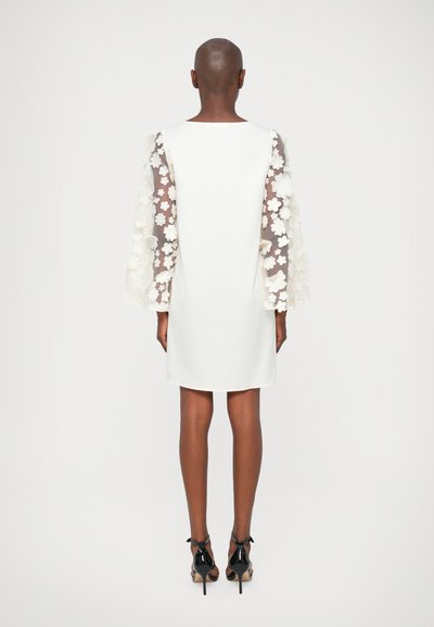 Vestido blanco con mangas transparentes de patrón floral, silueta recta y escote redondo, combinado con zapatos de tacón negro.