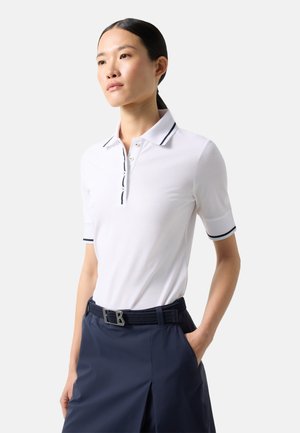 Woman wearing a white short-sleeve polo shirt with black trim and a navy skirt with a black belt, standing with one hand in pocket.