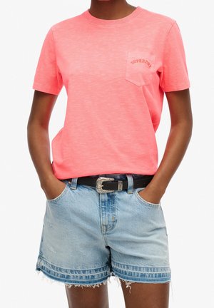 Person wearing coral pink short-sleeve T-shirt with chest pocket and light blue denim shorts with black belt, hands in pockets.