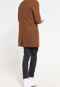 Cappotto lungo marrone con taglio diritto e spacco posteriore, abbinato a pantaloni scuri e sneakers bianche, visto da dietro su uno sfondo chiaro.