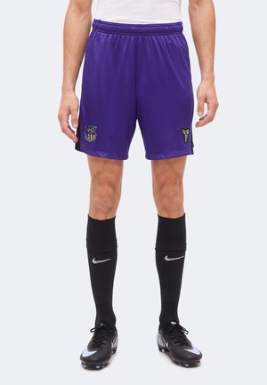 Homme portant un short de sport violet avec les logos du FC Barcelone et de Kobe Bryant, des chaussettes Nike noires et des chaussures de football noires.