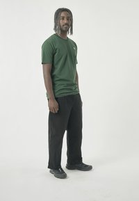 Dunkelgrünes T-Shirt mit kurzen Ärmeln und kleinem Logo, kombiniert mit lockeren schwarzen Hosen und schwarzen Turnschuhen; minimalistisches Design; einfarbige Farben.