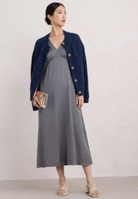 Abito grigio con pois, indossato sotto un cardigan in blu scuro con texture e grandi bottoni, abbinato a sandali beige con cinturini e una pochette.