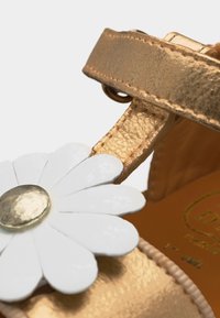 Κοντινό πλάνο λουριού χρυσού σανδαλιού με διακοσμητικό σε σχήμα λευκού χαμομηλιού και μεταλλικό χρυσό κεντρικό κουμπί.