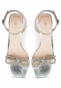 Silver high-heeled sandals with a square toe, adorned with a rhinestone-embellished bow and a thin ankle strap. Smooth leather texture.