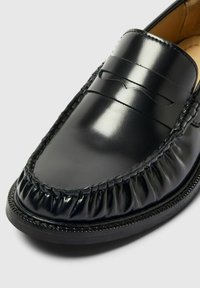 Svart lær slip-on loafer med glanset finish, med en rynket design ved tåen og horisontale sømmer over vamps.