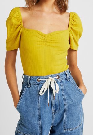 Vrouw draagt een geel geribbeld shirt met pofmouwen en een vierkante halslijn, gecombineerd met een hoge taille blauwe spijkerbroek met een witte trekkoord.