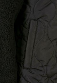Čierna bunda s textúrovaným fleecovým exteriérom a hladkým prešívaným podšívkou. Obsahuje malú vertikálnu kapsu vo vnútri.