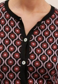 Cárdigan de punto con un patrón floral en negro y rosa, con acentos rojos. Tiene un cierre de botones en la parte delantera con dos botones de color claro.