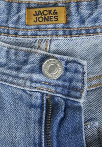 Jean en denim bleu clair avec un bouton en laiton, fermeture éclair et coutures beige. Étiquette "JACK & JONES" en fil doré cousue à la taille.