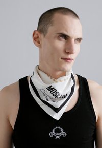 Witte zijden sjaal met zwarte rand waarop de tekst "Moschino" staat; gedragen om de hals over een zwart tanktop. Gladde textuur, rechthoekige vorm.