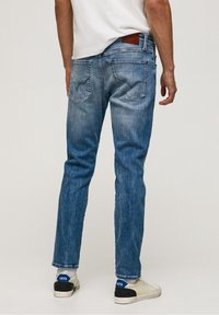 Hombre vestido con jeans azules desgastados, zapatillas blancas con detalles azules y una camiseta blanca, de pie de espaldas a la cámara frente a un fondo liso.