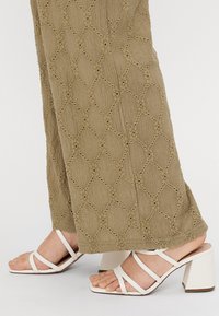 Λευκές ψηλοτάκουνες σανδάλες με λουράκια και τετράγωνη καμπύλη. Το πάνω μέρος διαθέτει πολλαπλά λεπτά λουράκια. Το φόντο δείχνει υφασμάτινη-textured ελιάς πράσινο.