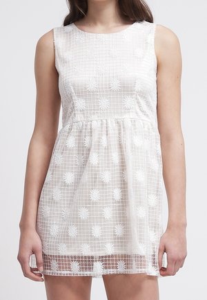 Mouwloze witte mini-jurk met vierkante halslijn, ruitpatroon en bloemenborduursel, gedragen door een persoon die met ontspannen armen staat.