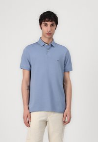 Polo shirt azzurro chiaro con colletto, maniche corte e tre bottoni. Piccolo logo sul lato sinistro del petto. Indossato con pantaloni di colore chiaro.
