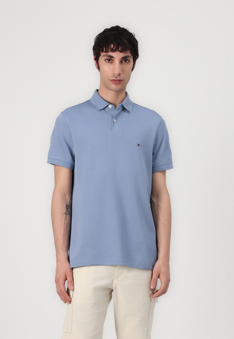 Polo shirt azzurro chiaro con colletto, maniche corte e tre bottoni. Piccolo logo sul lato sinistro del petto. Indossato con pantaloni di colore chiaro.
