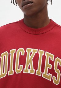 Felpa rossa con collare a costine, caratterizzata da un grande testo ricamato "DICKIES" in oro e bianco. Tessuto morbido con una finitura texturizzata.