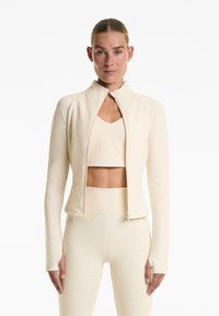 Giacca beige con zip e colletto alto, maniche lunghe e texture a coste, abbinata a un crop top coordinato e leggins aderenti.