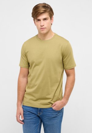 Young man with light brown hair wearing a plain olive green T-shirt and blue jeans, standing with one hand in pocket against white background.