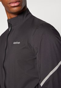 Schwarze Radjacke mit hohem Kragen, Frontreißverschluss und reflektierenden Akzenten an den Schultern. Leicht und mit glatter Textur für Komfort.