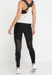 Mallas deportivas negras con paneles de malla, que presentan texto blanco en una pierna. Combinadas con una camiseta sin mangas de color claro y un sujetador deportivo negro.