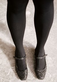 Zapatos negros tipo Mary Jane con textura brillante, puntera cuadrada y tira en el tobillo, combinados con medias opacas negras. Brillo sutil y diseño robusto.