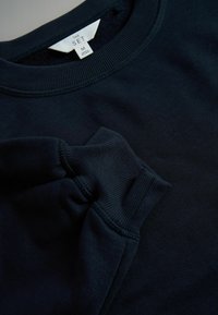 Tumsummā zila kapuce ar ribotām apkaklēm un piedurknēm. Mīksta auduma ar gludu tekstūru, etiķete iemontēta apkakles iekšpusē. Vidēja izmēra.