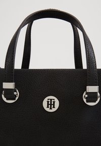 Borsa a tracolla in pelle nera con superficie testurizzata, due manici corti e dettagli in metallo argentato, caratterizzata da un logo circolare al centro.