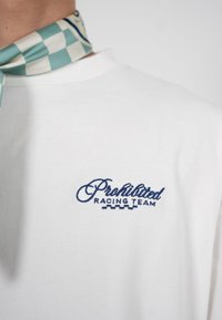 Bela srajca z modrim vezenjem "Prohibited Racing Team" in zeleno-belo karo ruto, zavezano okrog vratu.