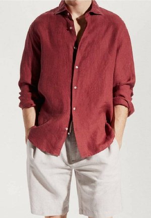 Mann trägt ein langärmliges, aufgeknöpftes rotes Hemd mit hochgekrempelten Ärmeln und hellbeige Shorts, die Hände in den Taschen, steht vor einem einfarbigen Hintergrund.