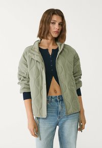 Hellgrüne gesteppte Jacke mit hohem Kragen, Reißverschluss vorne und großen Seitentaschen. Über einem dunkelblauen, gerippten Oberteil und hochtaillierten Jeans getragen.