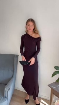 Vestido roxo com ombros à mostra em tecido elástico, silhueta ajustada, mangas longas, drapeado lateral, combinado com uma bolsa preta e sapatos de salto agudo.
