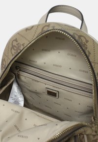 Interno di uno zaino beige Guess, con più scomparti, fodera branding e una tasca con zip per un trasporto sicuro.