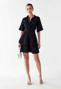 Vestido negro de camisa con mangas cortas, delantero anudado y dobladillo en capas, hecho de una tela texturizada. Combinado con un bolso blanco y sandalias.