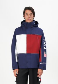 Mand iført en navy, hvid og rød hættejakke med "USA", et amerikansk flag og "Sail GP Team" logoer på højre ærme.
