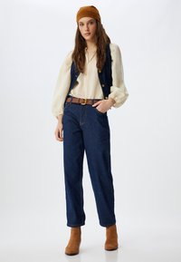Chaleco de denim con botones de latón sobre una blusa color crema, combinado con jeans azul oscuro y un cinturón de cuero marrón. Botines de ante marrón completan el atuendo.