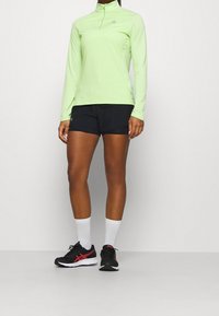 Ljust grön långärmad tröja med halvdragkedja, ihop med svarta shorts. Svart och röd träningsskor och vita strumpor fulländar outfiten.