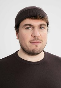 Bruine gebreide beanie met ribbelstructuur, gedragen bovenop een donkere trui met een ronde halslijn. Heeft een neutrale achtergrond en warme verlichting.