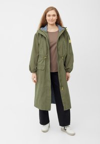 Giacca lunga verde oliva con vita regolabile, tasche frontali e fodera del cappuccio a righe blu. Indossata sopra una camicia marrone e pantaloni neri.