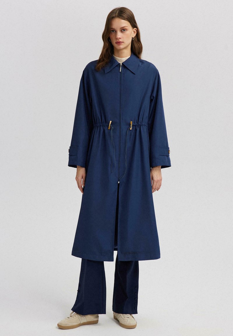 Tamno plavi dugi kaput sa prednjim patentnim zatvaračem, kabelom za podešavanje u struku i širokim manžetama. Ispod nosi par odgovarajućih tamnoplavi hlača.