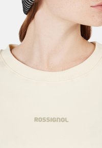 Sudadera color crema con cuello acanalado, que presenta el texto "ROSSIGNOL" en un gris sutil. La tela parece suave con una textura lisa.